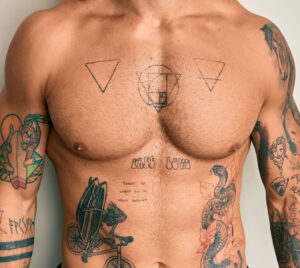Torso nudo di giovane uomo dalla pelle molto chiara, con tatuaggi su braccia petto e pancia.