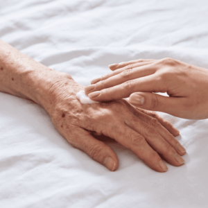 Mano di anziana donna sdraiata a letto, su lenzuola bianche, a cui viene applicata della crema base