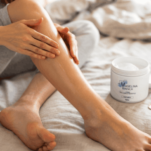 Primo piano delle gambe di una donna, seduta su un letto con delle lenzuola chiare, che si spalma un sottile strato di Vaselina Bianca sulle ginocchia